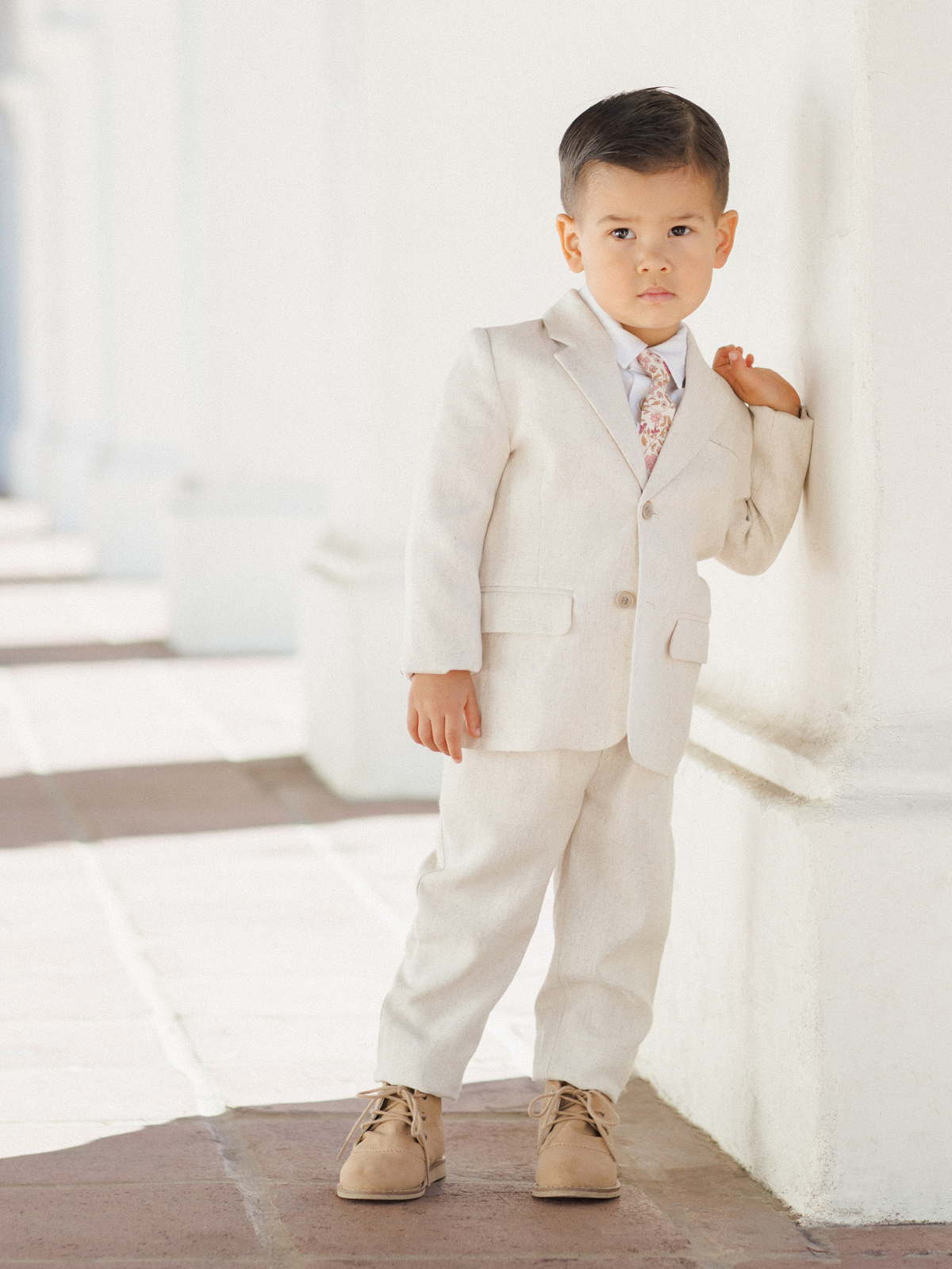 Young boy in a beige suit and shoes, posing against a light wall in a stylish, formal setting.