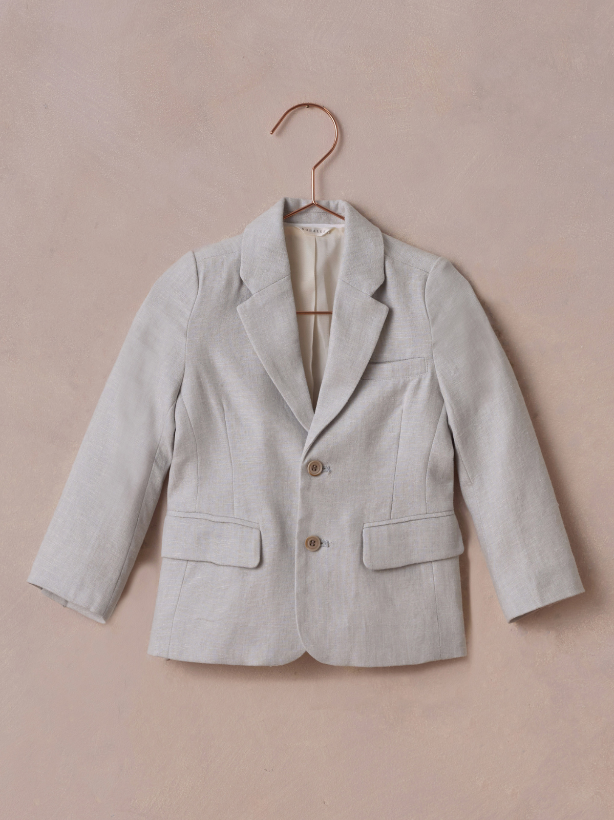 Light gray blazer hanging on a copper hanger against a textured pink background.