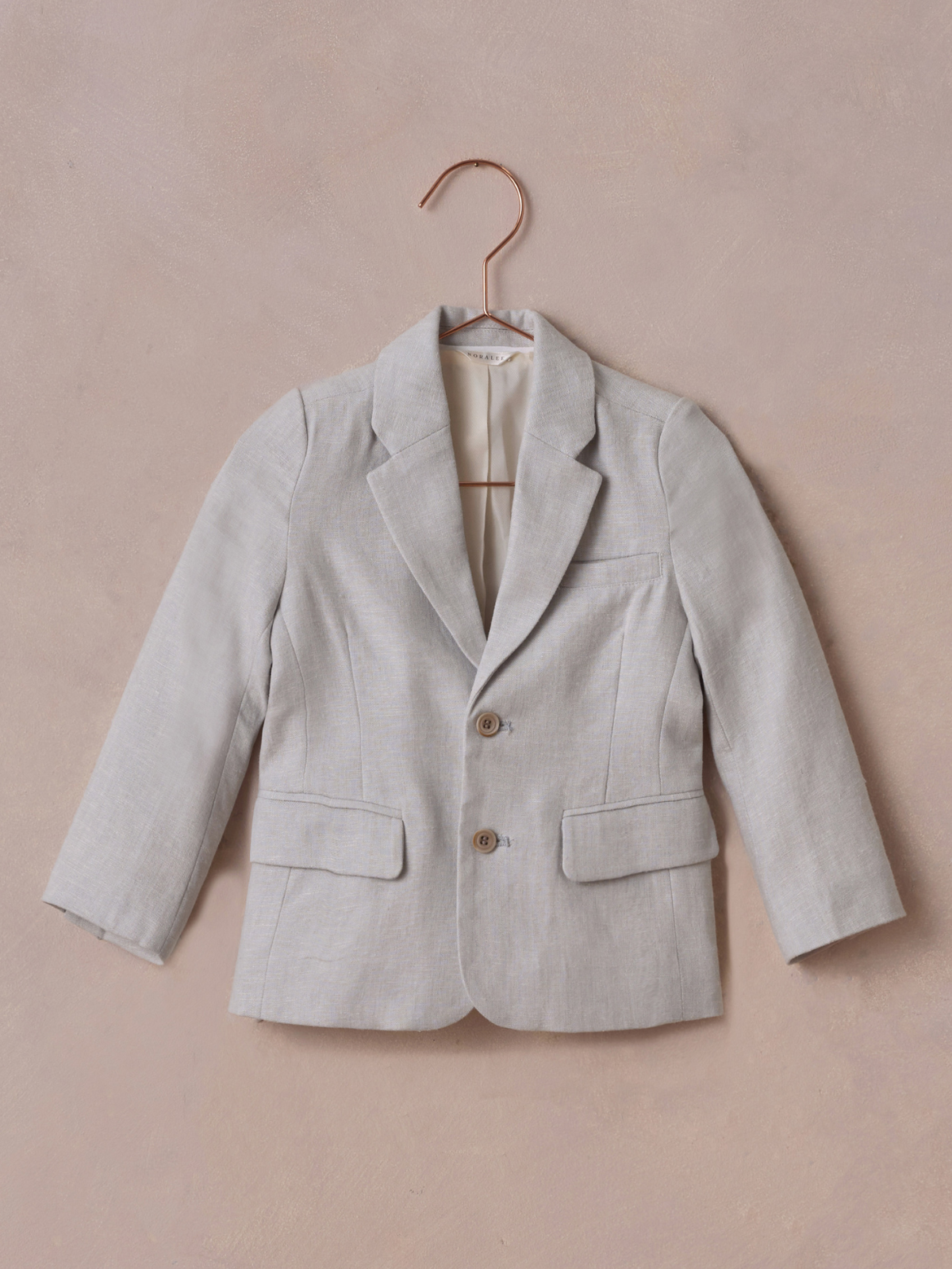 Light gray blazer hanging on a copper hanger against a textured pink background.