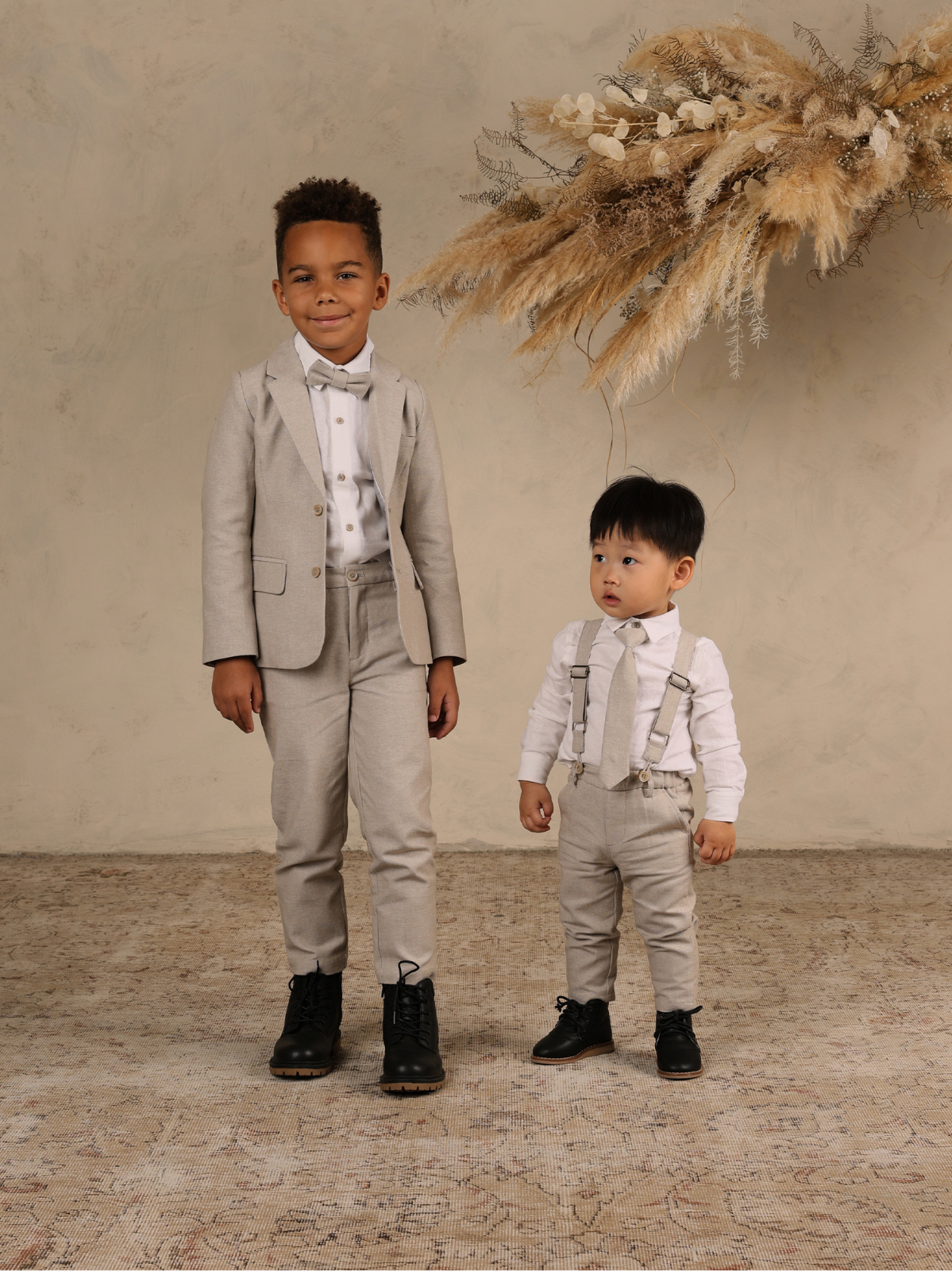 Two young boys in stylish suits pose for a photo, with a decorative backdrop of dried flowers.