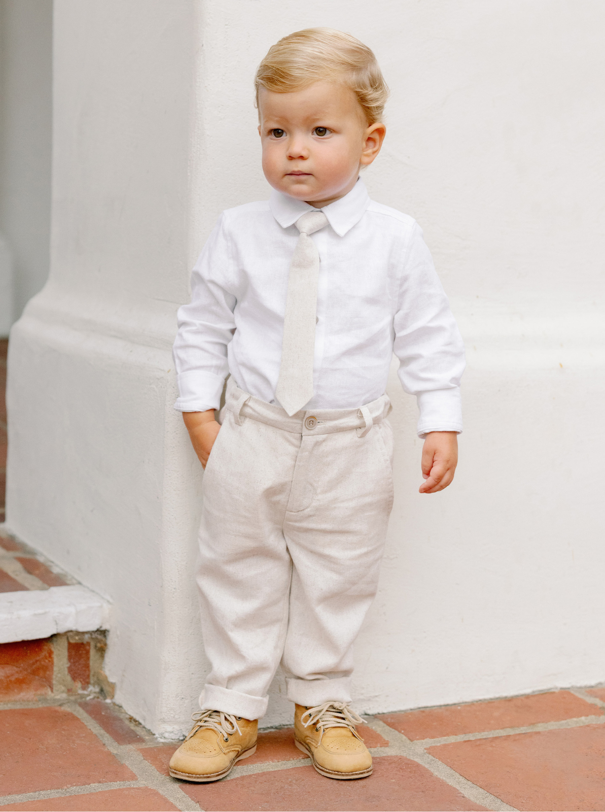 A young boy in a white shirt and beige trousers poses against a white wall, looking thoughtfully to the side.