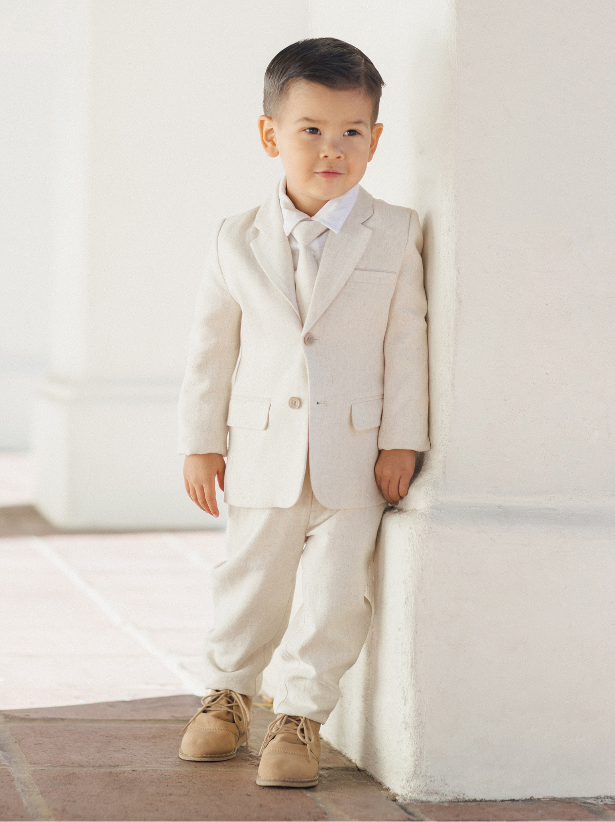 Young boy in a light suit and tie, standing by a wall with a confident expression.