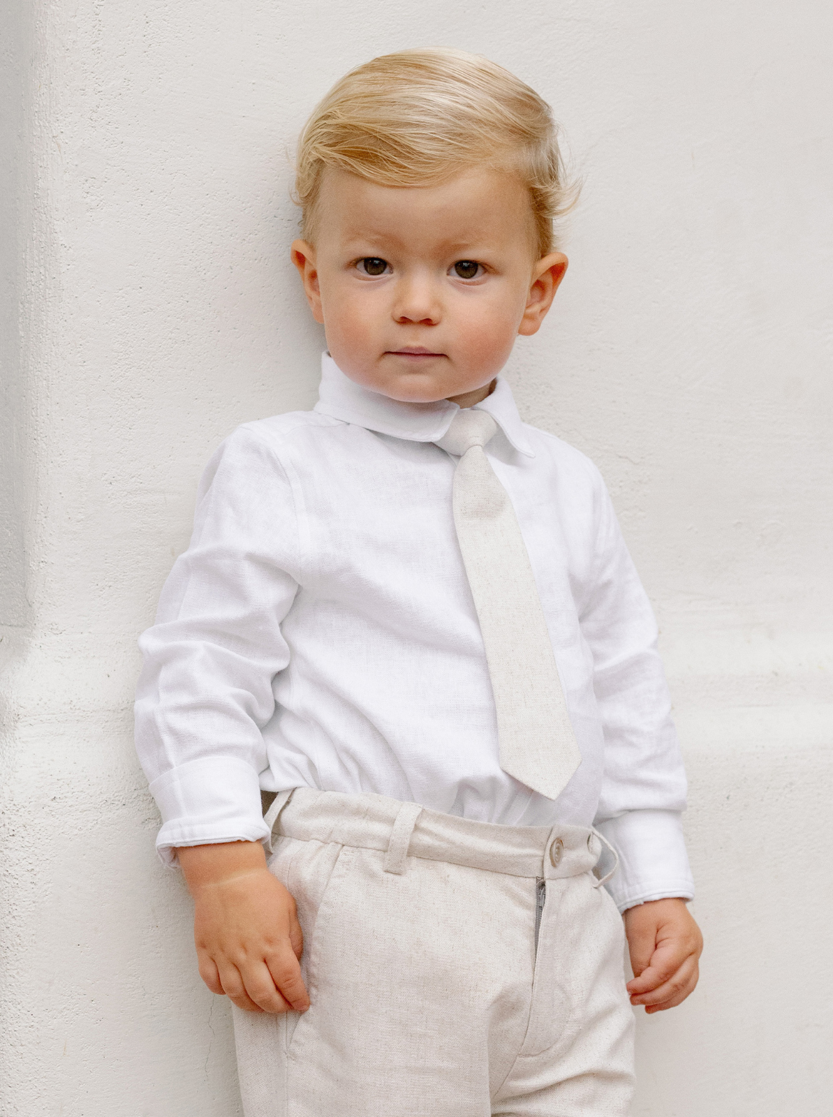 Young boy in formal white shirt and light-colored tie, standing against a textured white wall.