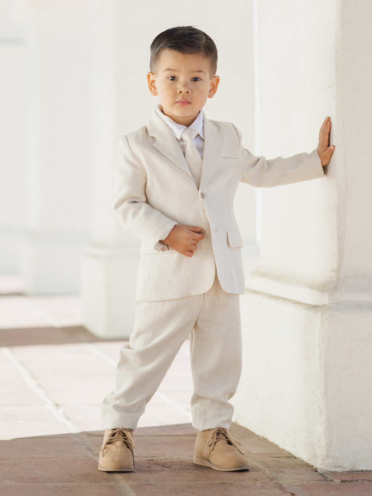 A young boy in a beige suit stands confidently against a white wall, wearing stylish shoes.