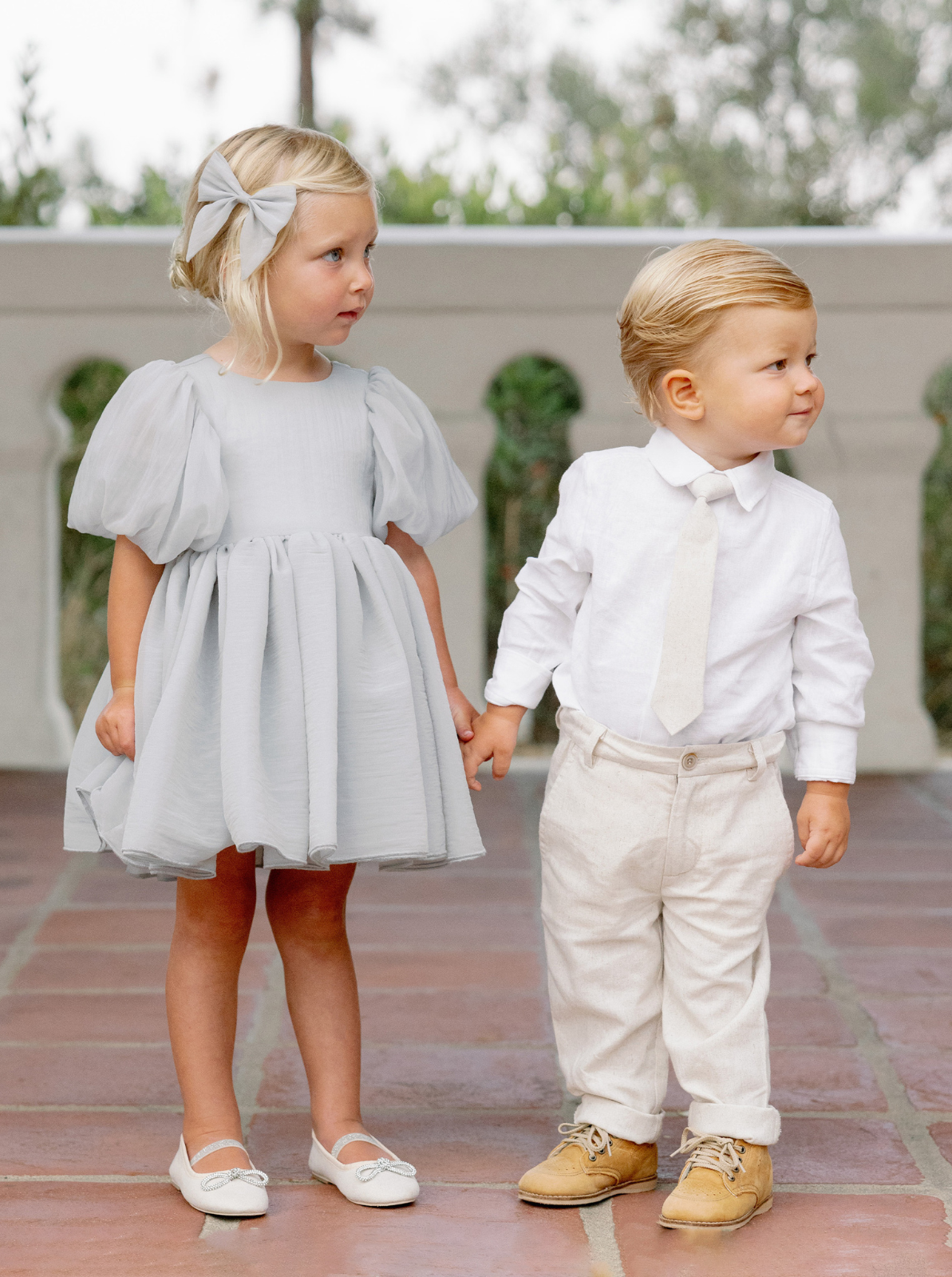 Two young children hold hands outdoors; a girl in a gray dress and a boy in a white shirt and light pants.