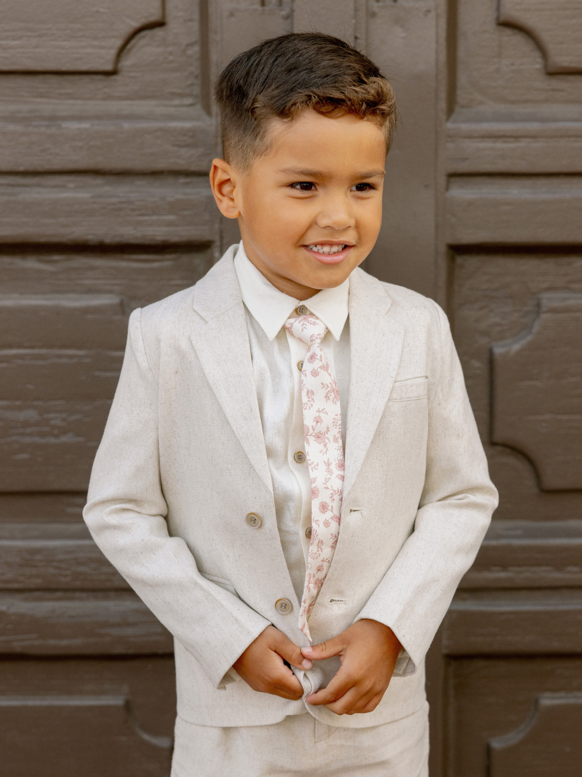 Smiling boy in a light suit and floral tie, standing against a wooden backdrop.