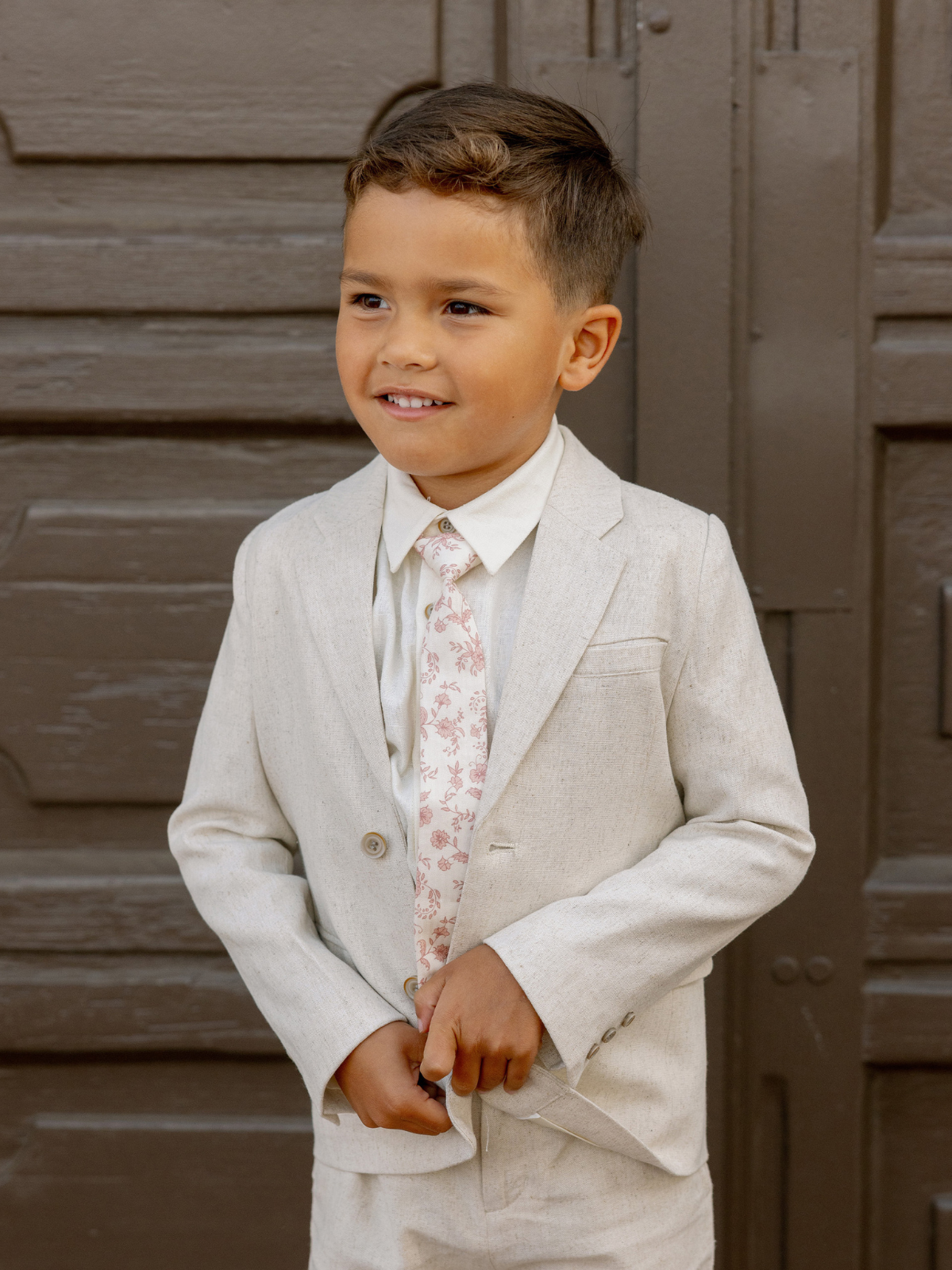 Young boy in a light gray suit and floral tie, smiling against a brown wooden backdrop.