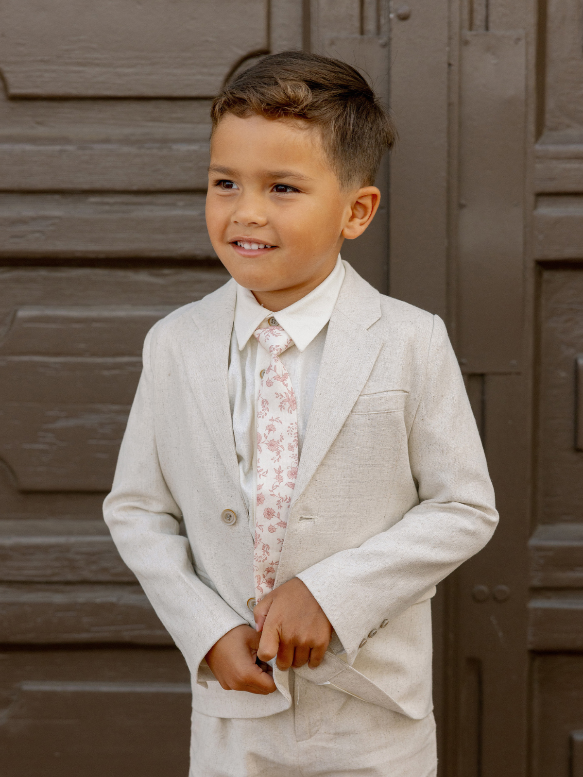 Young boy in a light gray suit and floral tie, smiling against a brown wooden backdrop.