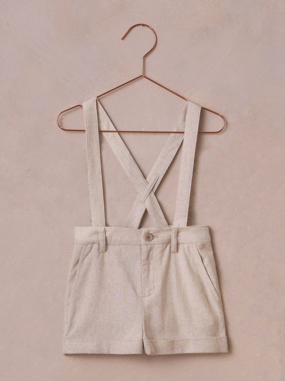 Beige shorts with cross-back suspenders hanging on a rose gold hanger against a soft pink background.
