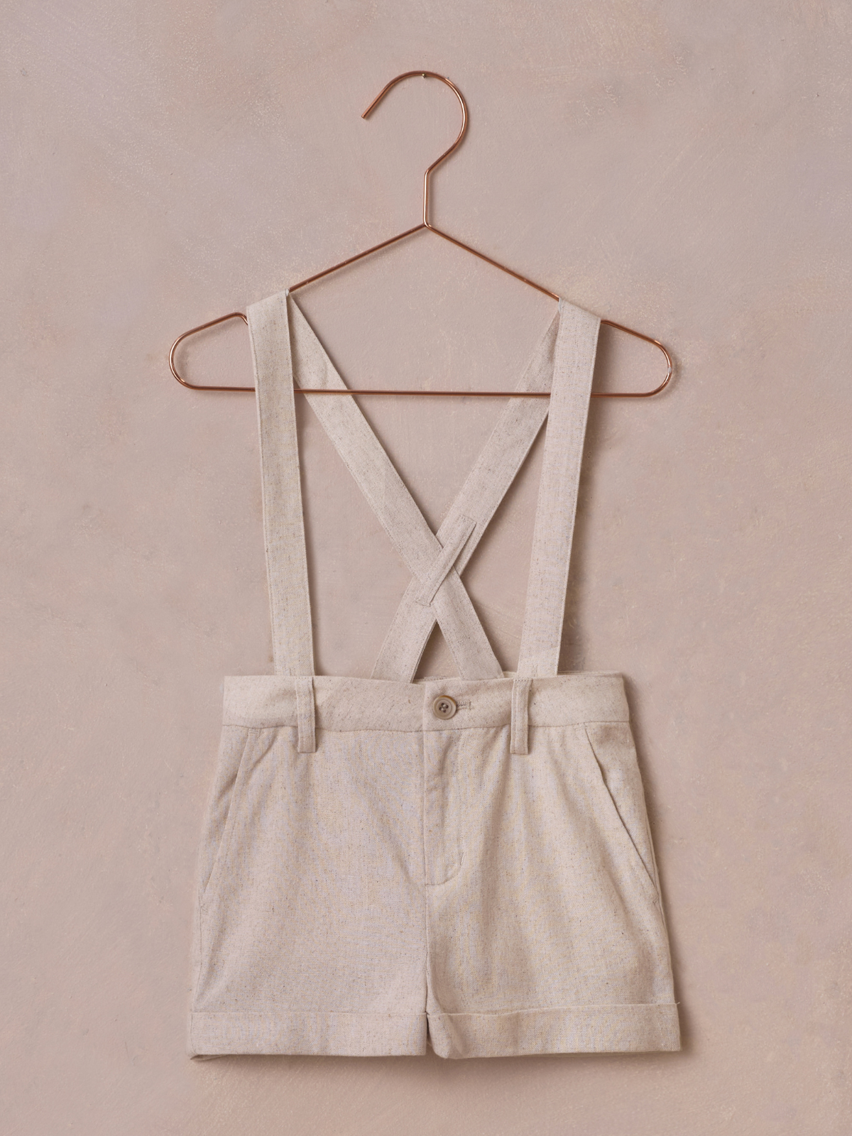 Beige shorts with cross-back suspenders hanging on a rose gold hanger against a soft pink background.
