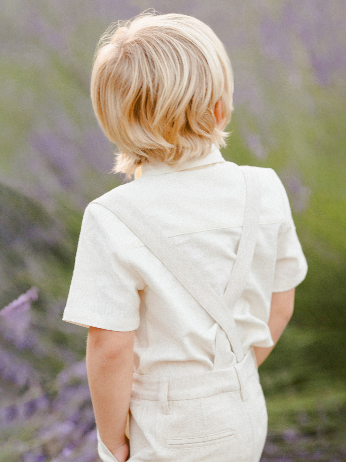 Child with blonde hair wearing light clothing, standing in a lavender field, facing away from the camera.