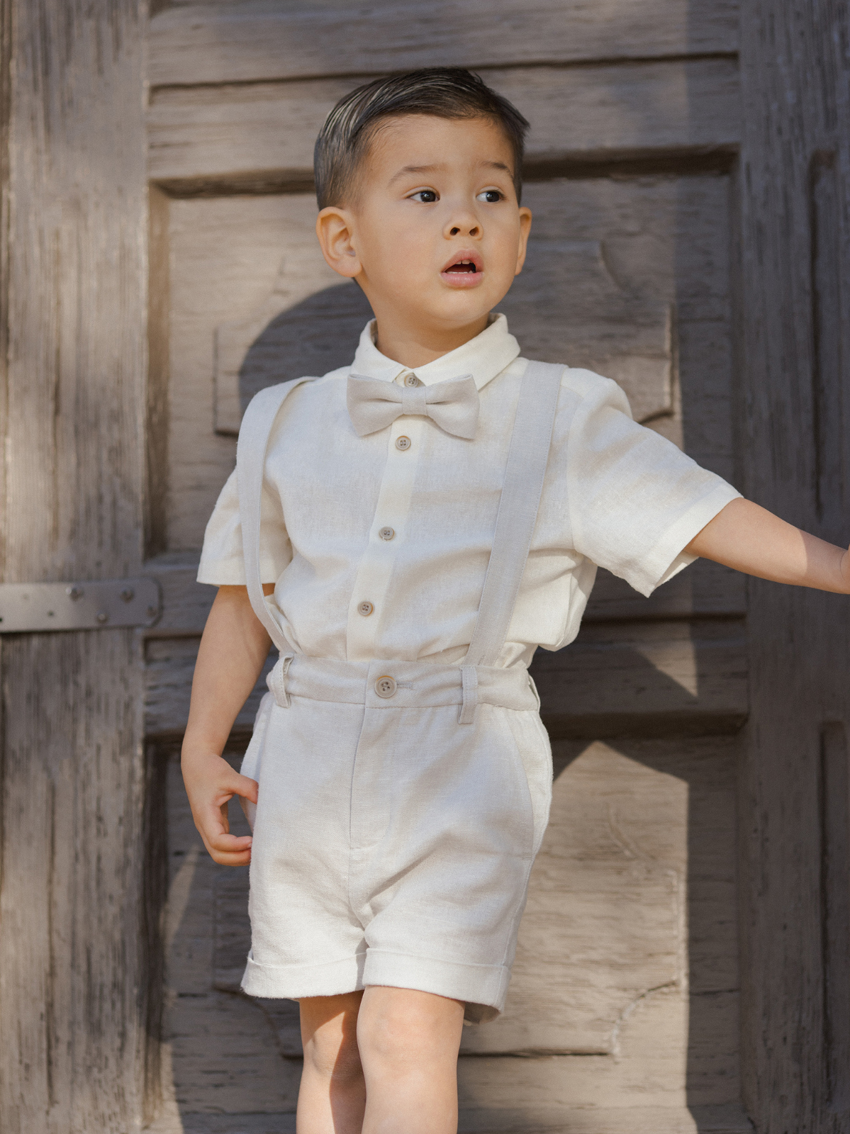 Child in a light-colored outfit with suspenders and a bow tie, standing in front of a wooden door.