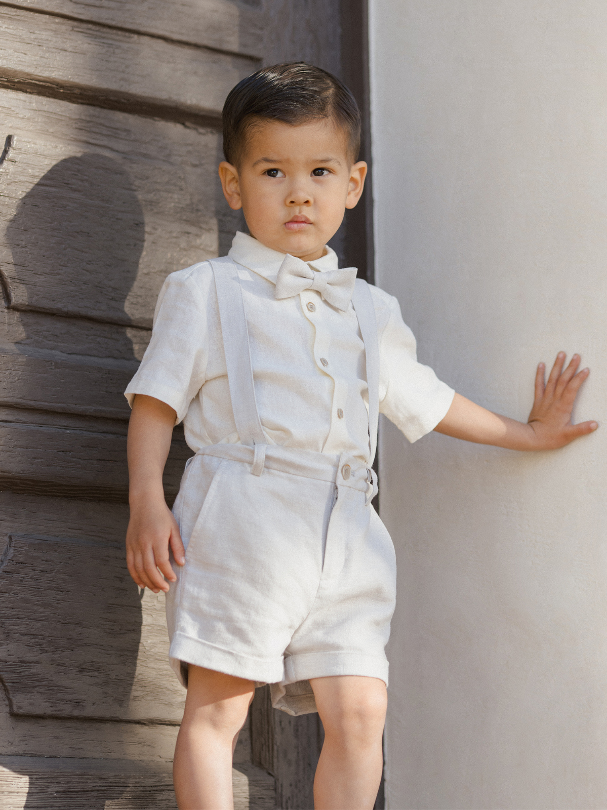 Young boy in cream outfit with bow tie and suspenders, standing against a rustic wall.
