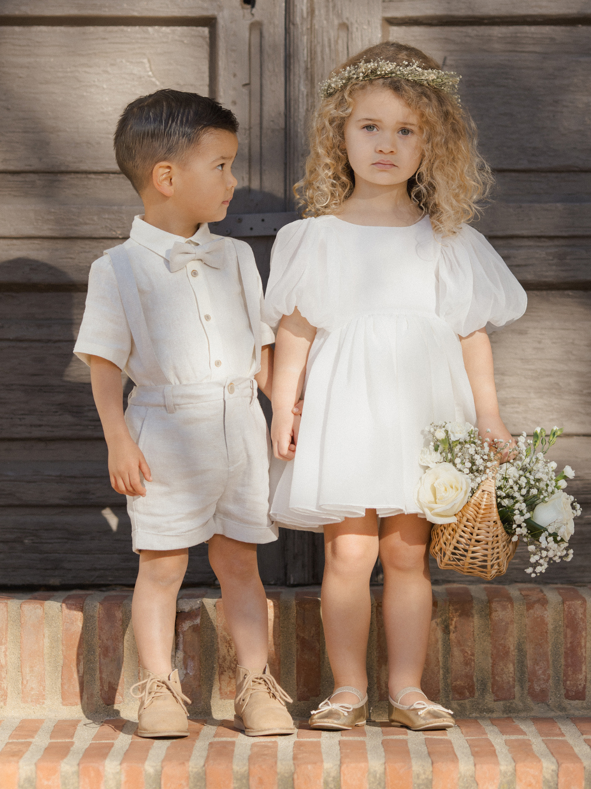 Two young children stand on brick steps; the girl in a white dress holds flowers, while the boy wears light-colored shorts and a bow tie.
