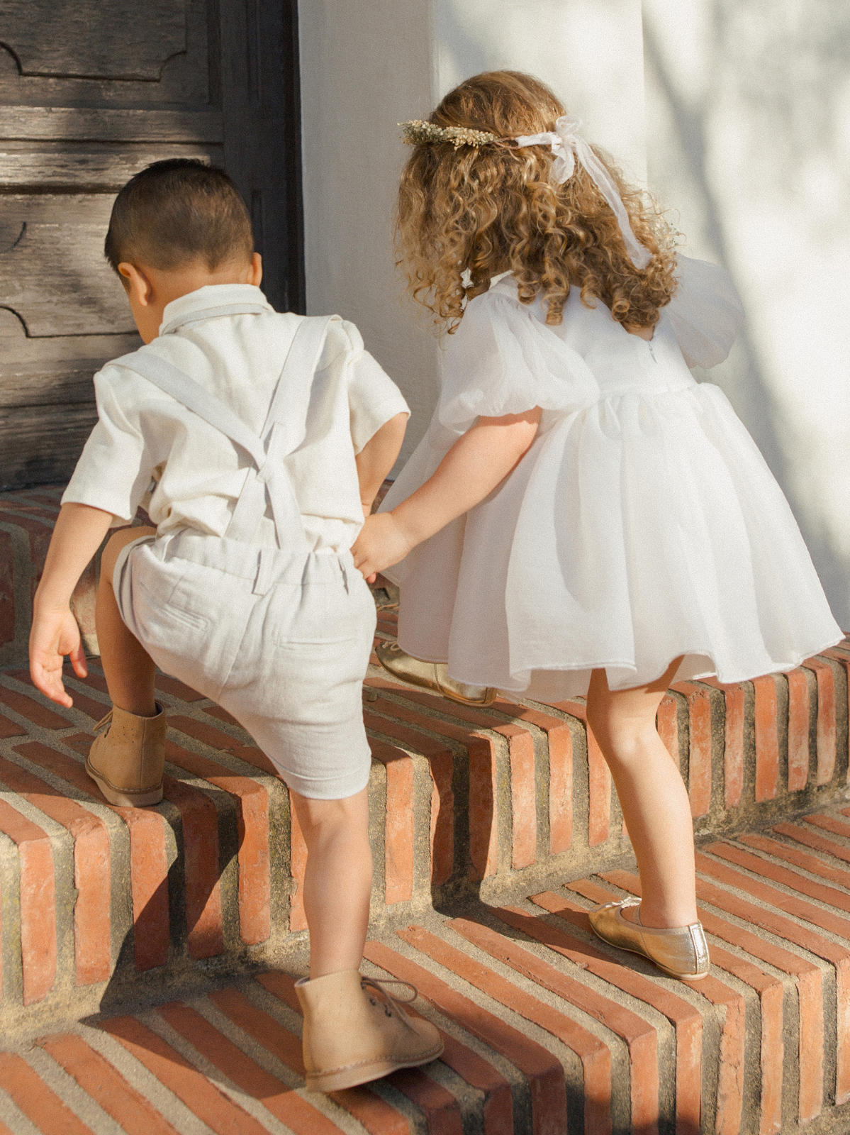 Two children in light outfits climbing brick steps, enjoying a playful moment together in the sunlight.