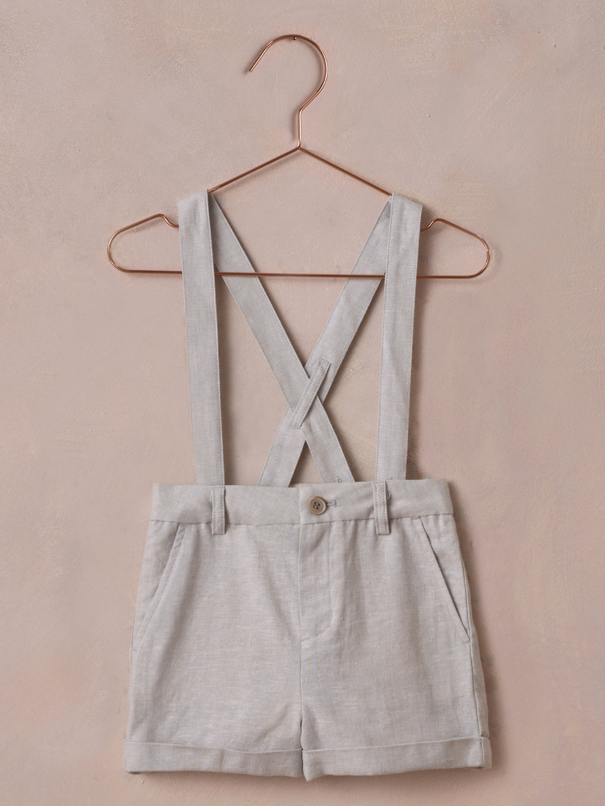 Gray shorts with crisscross suspenders hanging on a copper hanger against a light pink background.