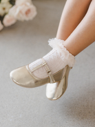 Gold ballet flats worn with white frilly socks on a child's feet, against a soft floral background.