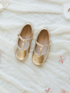 Gold toddler shoes with a strap, placed on light fabric alongside decorative bows.