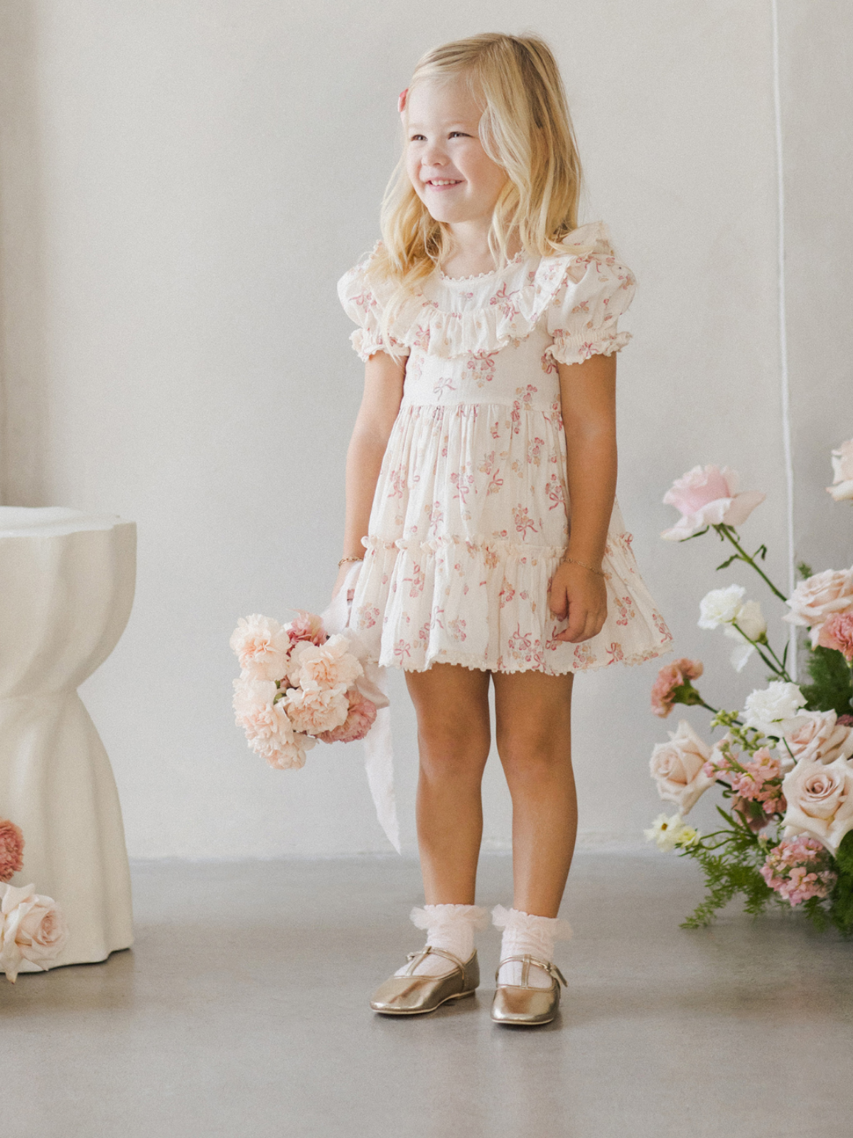 Smiling child in a floral dress holds a bouquet, surrounded by flowers in a bright, stylish setting.