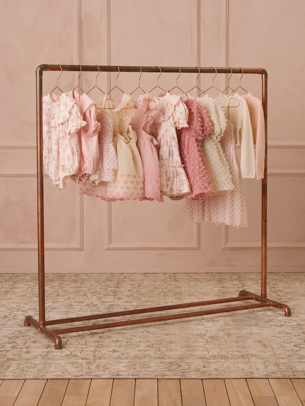 Pastel dresses hanging on a copper rack against a soft pink backdrop, showcasing an array of delicate designs.