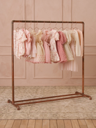 Pastel dresses hanging on a copper rack against a soft pink backdrop, showcasing an array of delicate designs.