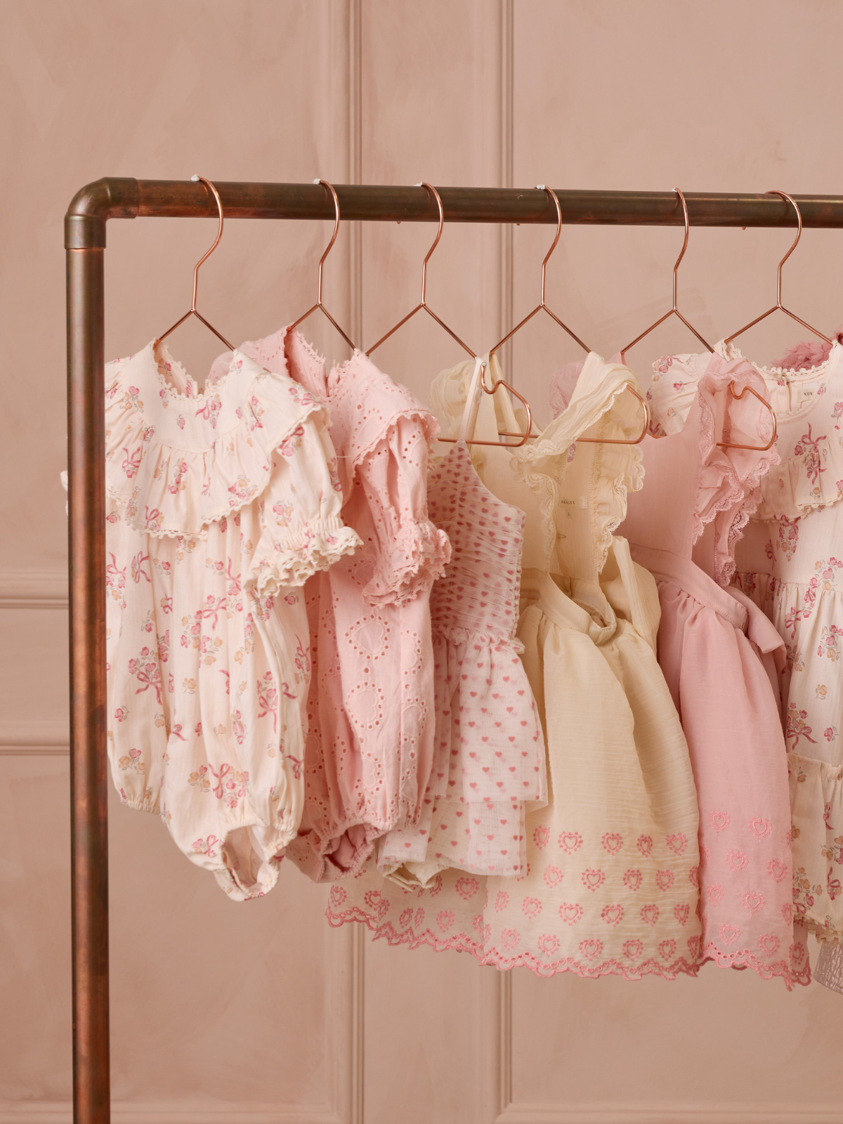 Stylish pink children's dresses hanging on a copper rack against a soft pink background.