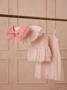 Three cute children's dresses hang on rose gold hangers against a soft pink background.