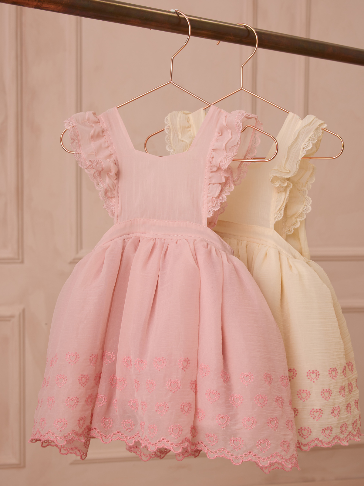 Two cute children's dresses hanging: one pink with ruffles, the other yellow, both with delicate embroidered details.