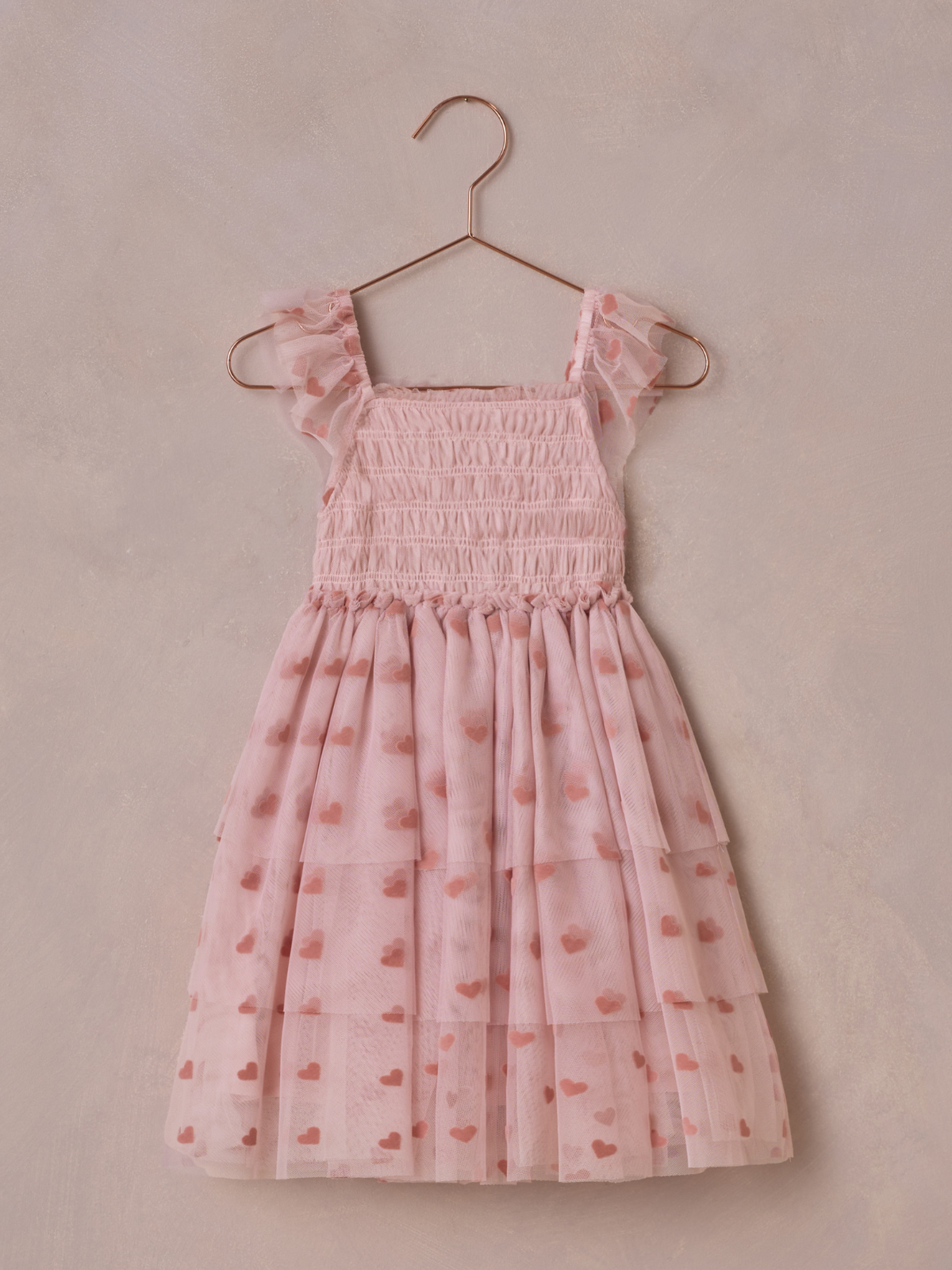 Light pink dress with heart patterns, ruffled straps, and layered skirt, displayed on a copper hanger against a neutral background.
