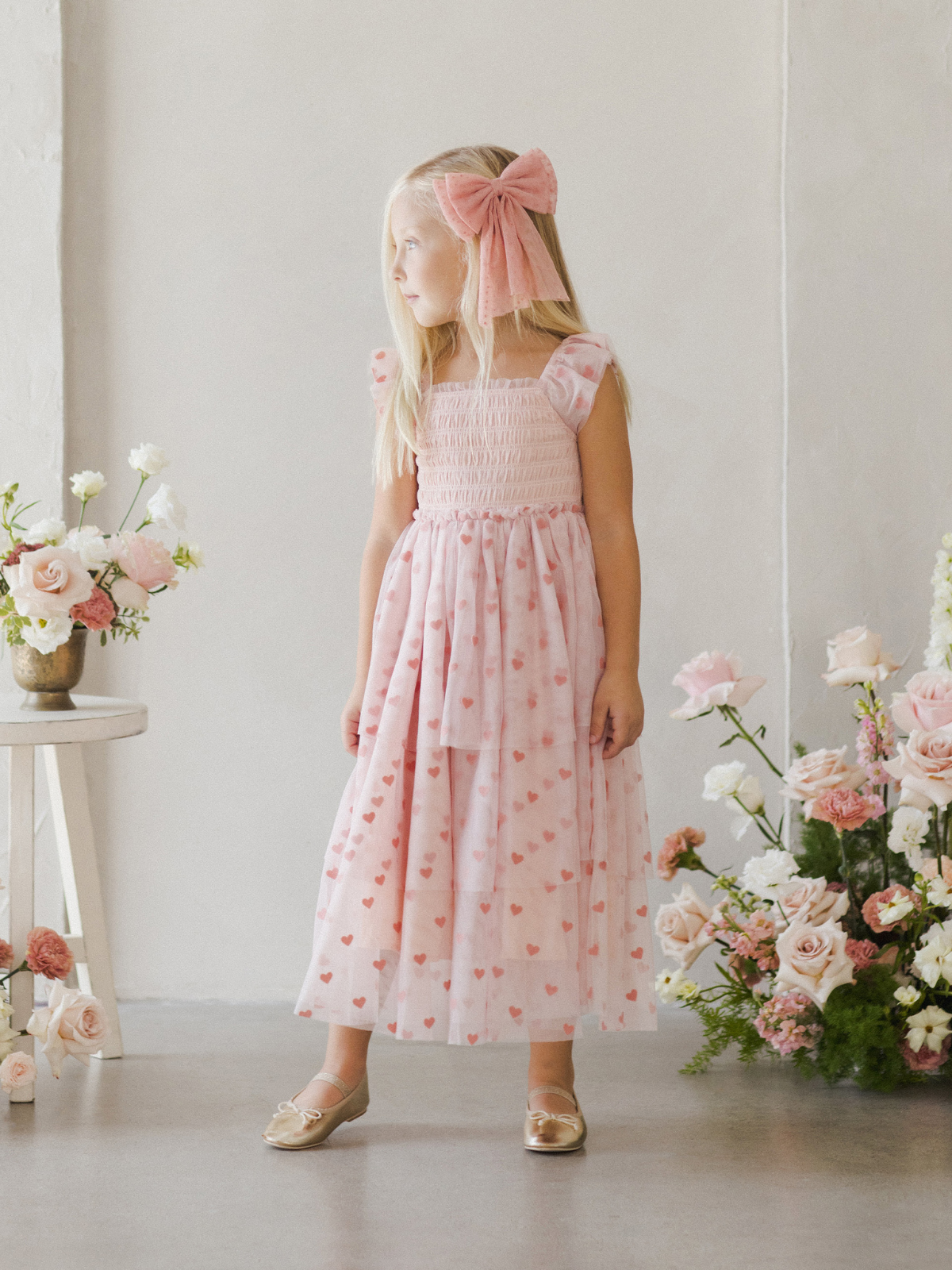 Girl in a pink heart-patterned dress stands by a flower arrangement in a bright, minimalist room.