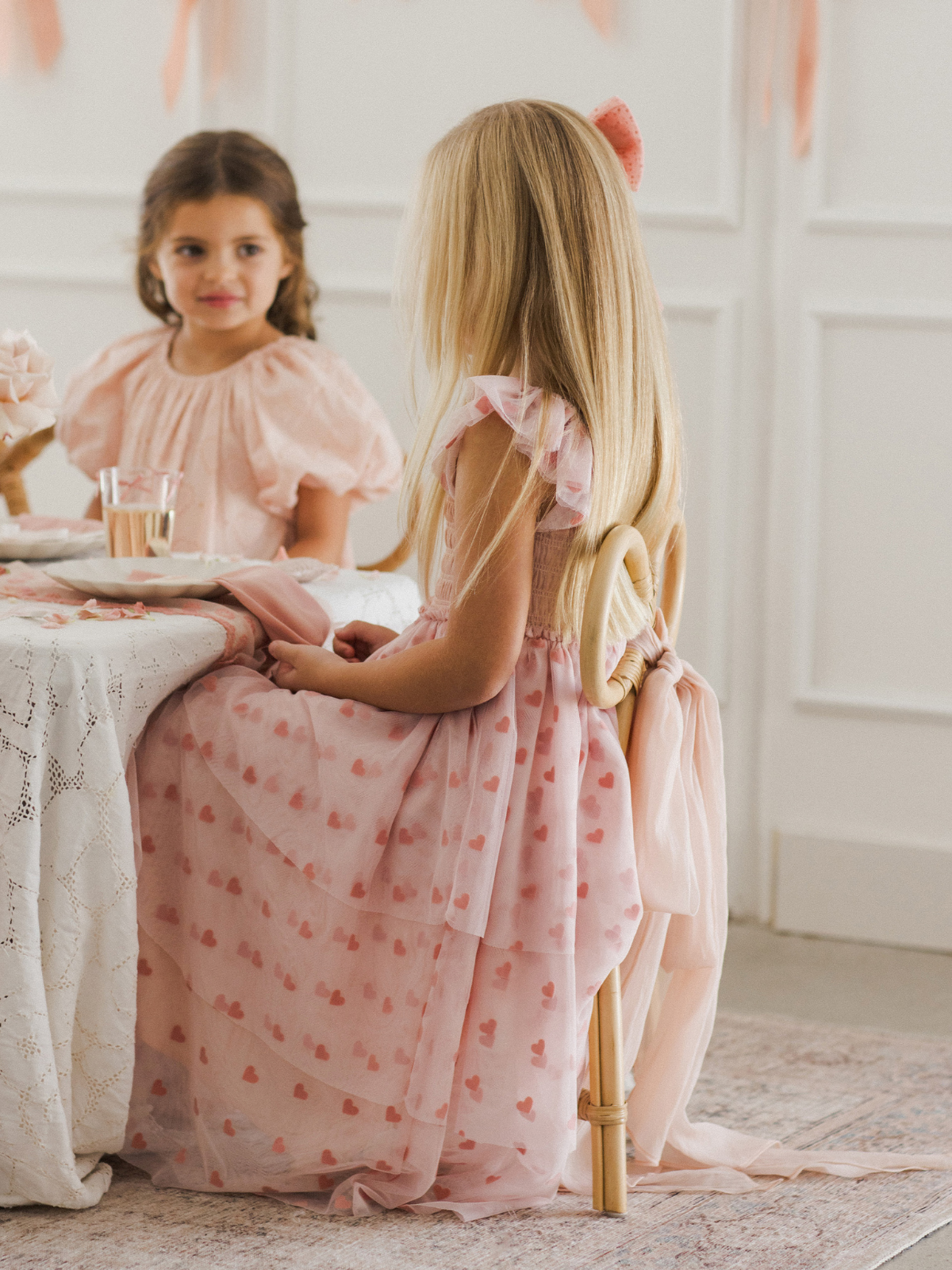 Two girls in pink dresses sit at a table decorated with flowers, enjoying a playful moment together.