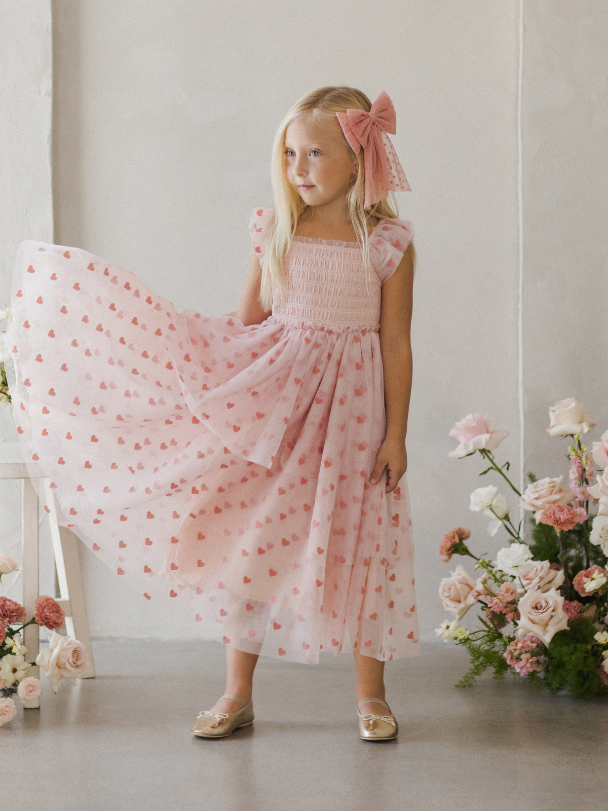 Girl in a pink heart-patterned dress with a large bow, standing among flowers in a softly lit room.