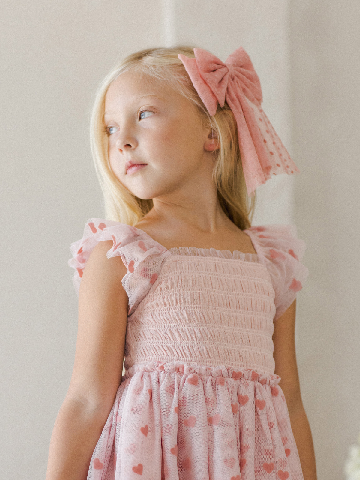 Young girl in a pink dress with heart patterns and a large pink bow, looking thoughtfully to the side.