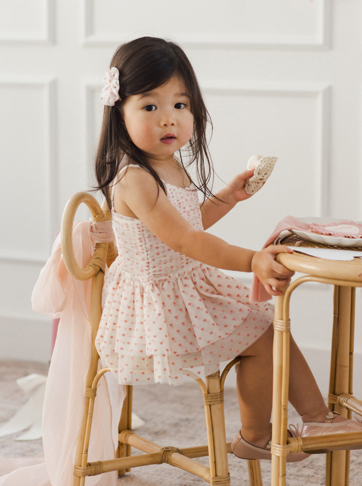 Young girl in a heart-patterned dress sitting at a table, holding a cookie, with soft lighting in a bright room.