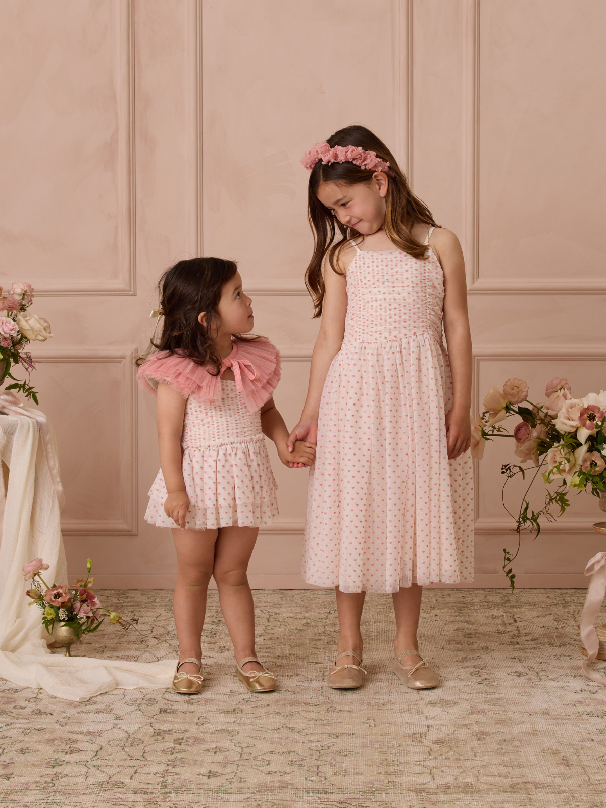 Two girls in pink dresses holding hands, posed in a soft, floral setting with a light pink background.