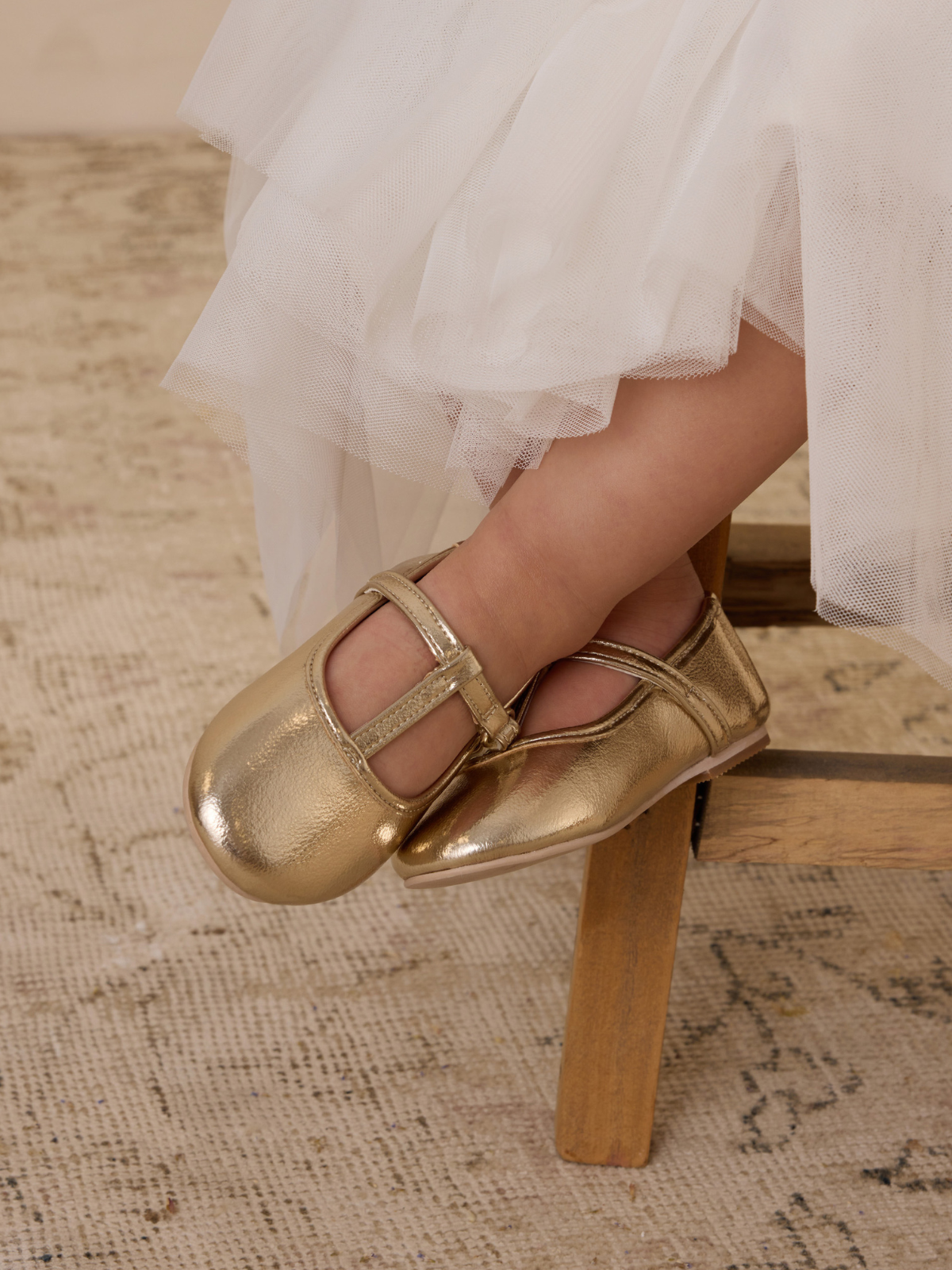 Gold ballet flats on a child's feet, resting on a wooden chair with tulle fabric in the background.