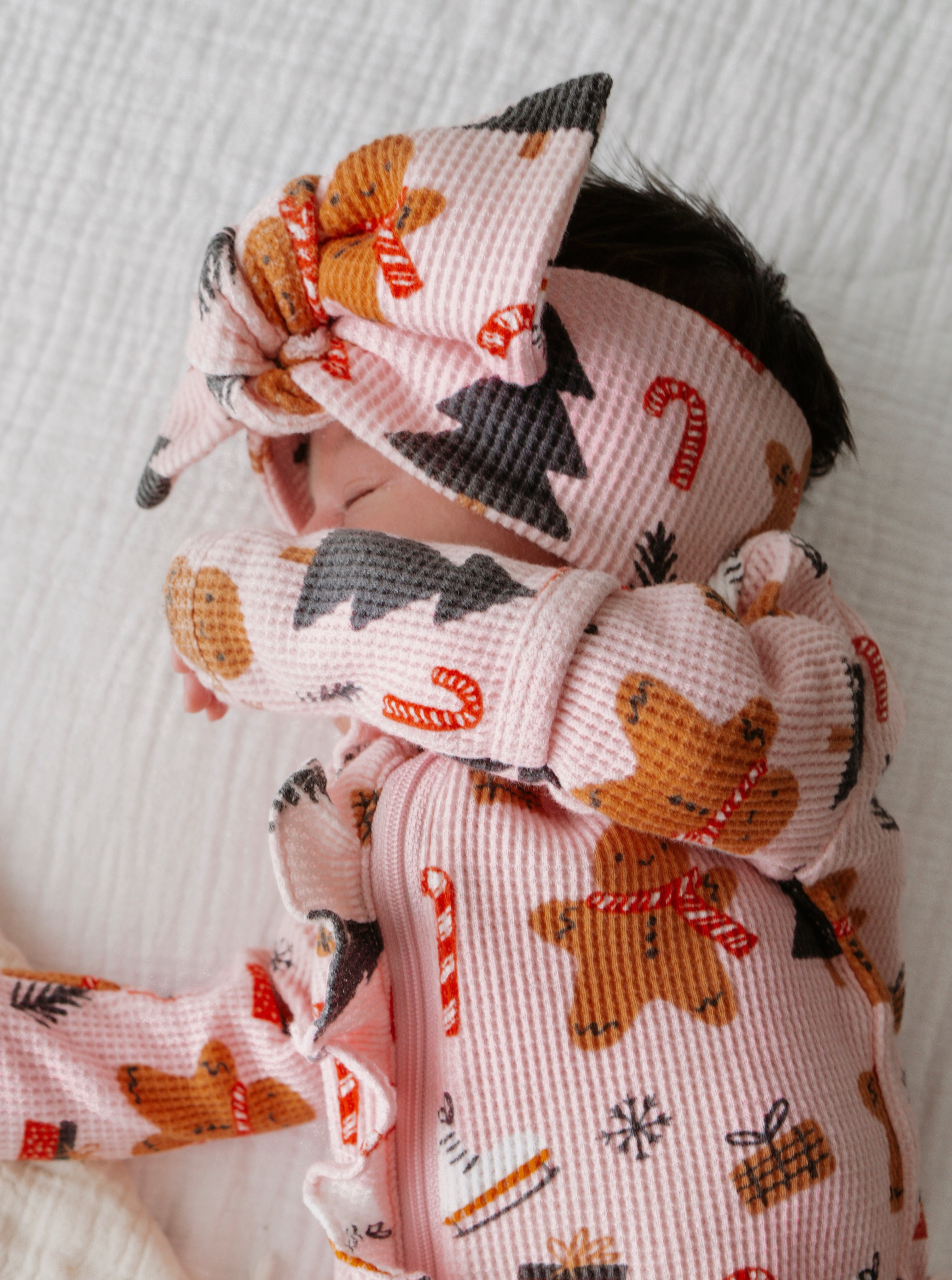 Baby in a festive pink outfit with gingerbread and candy cane patterns, resting on a textured white blanket.