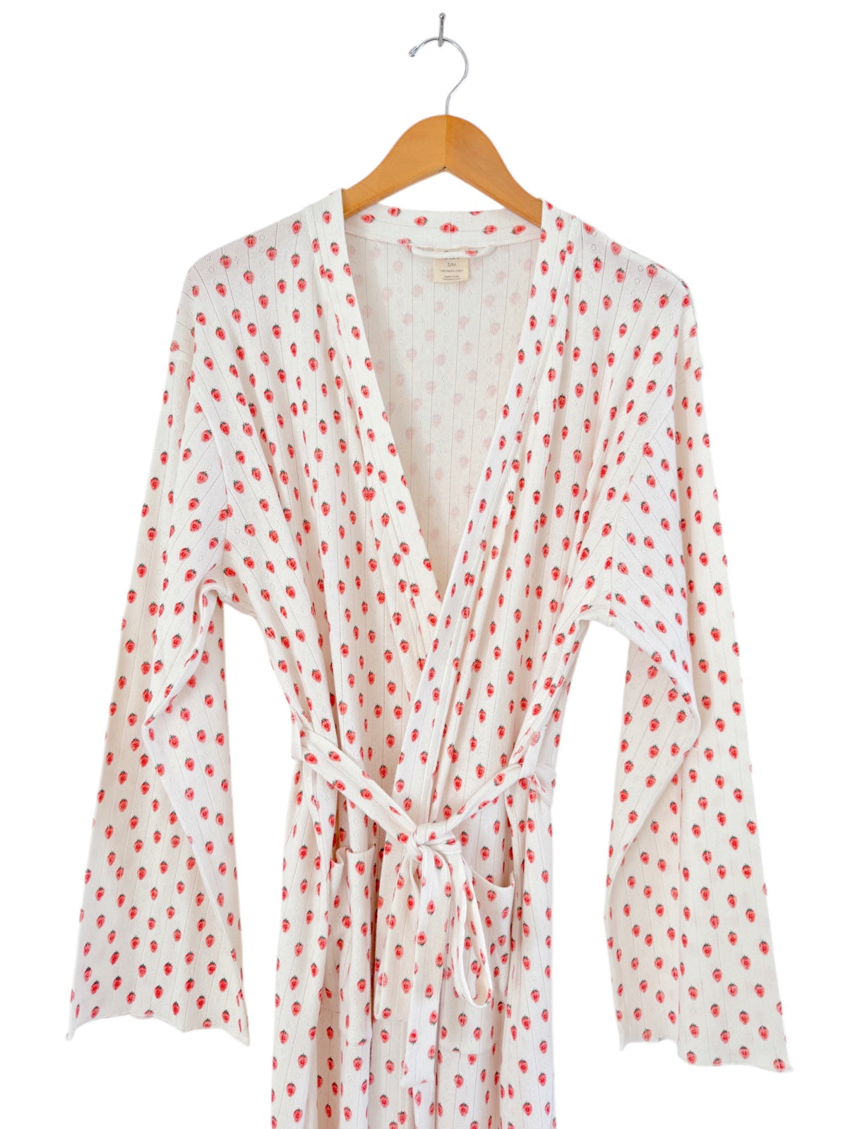 White robe with red strawberry print, hanging on a wooden hanger, displaying long sleeves and a tied waist.