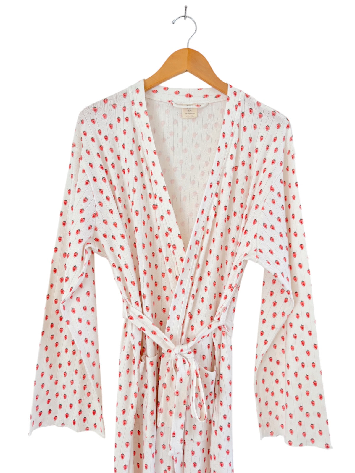 White robe with red strawberry print, hanging on a wooden hanger, displaying long sleeves and a tied waist.