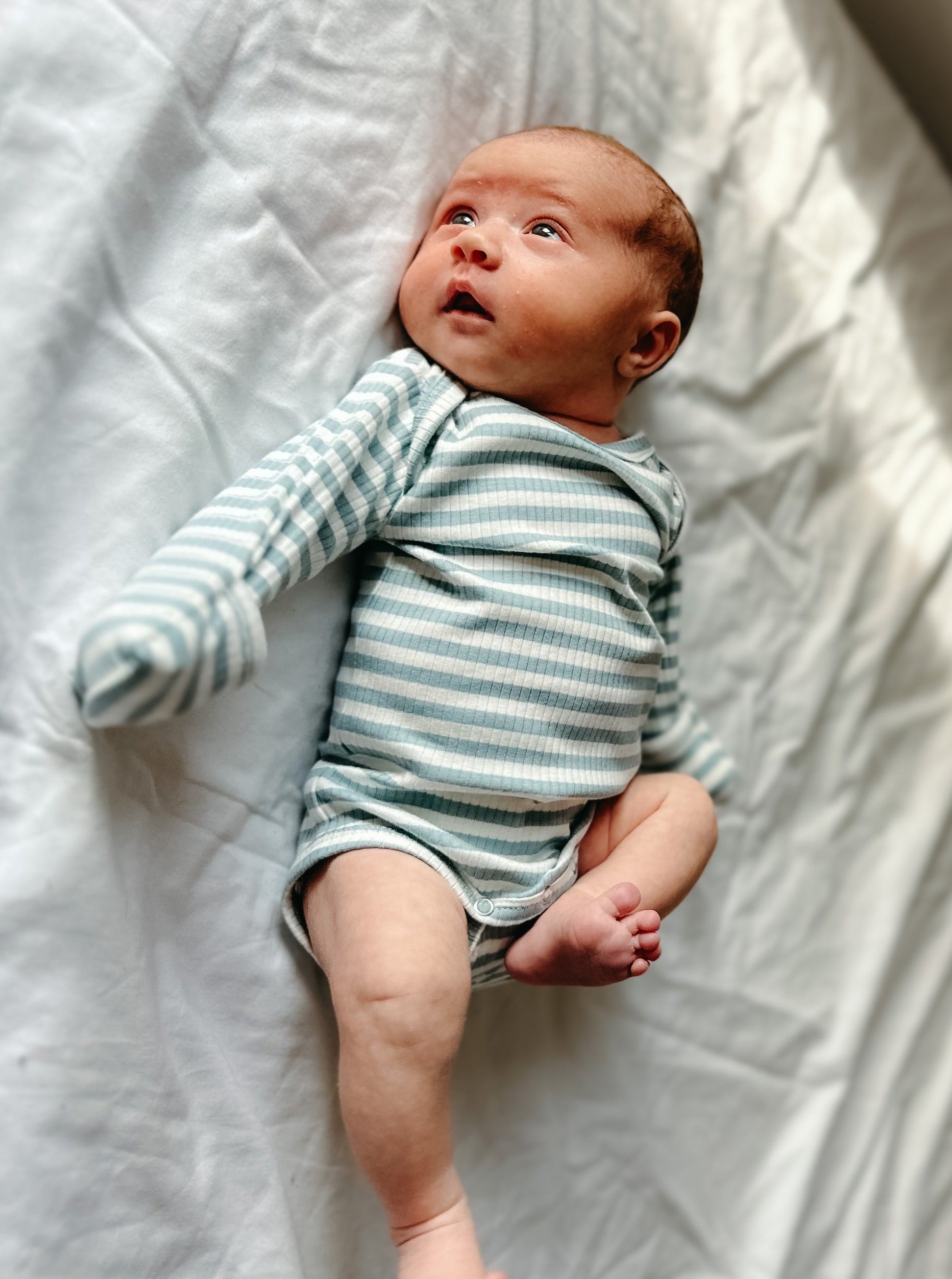Infant in a striped onesie lying on a white surface, looking up with wide eyes.