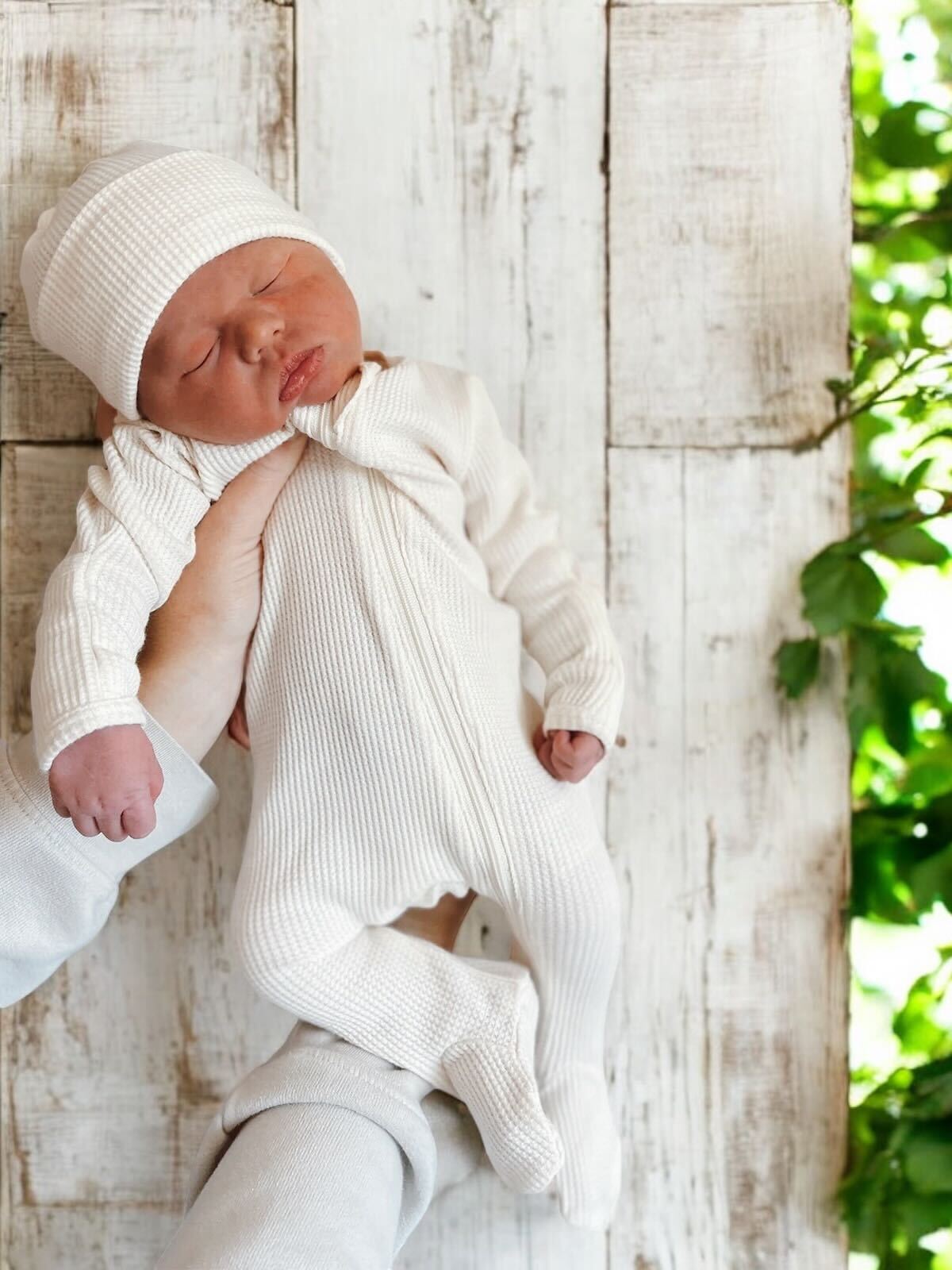 Newborn baby in white outfit and hat, being held against a rustic wooden background with greenery.