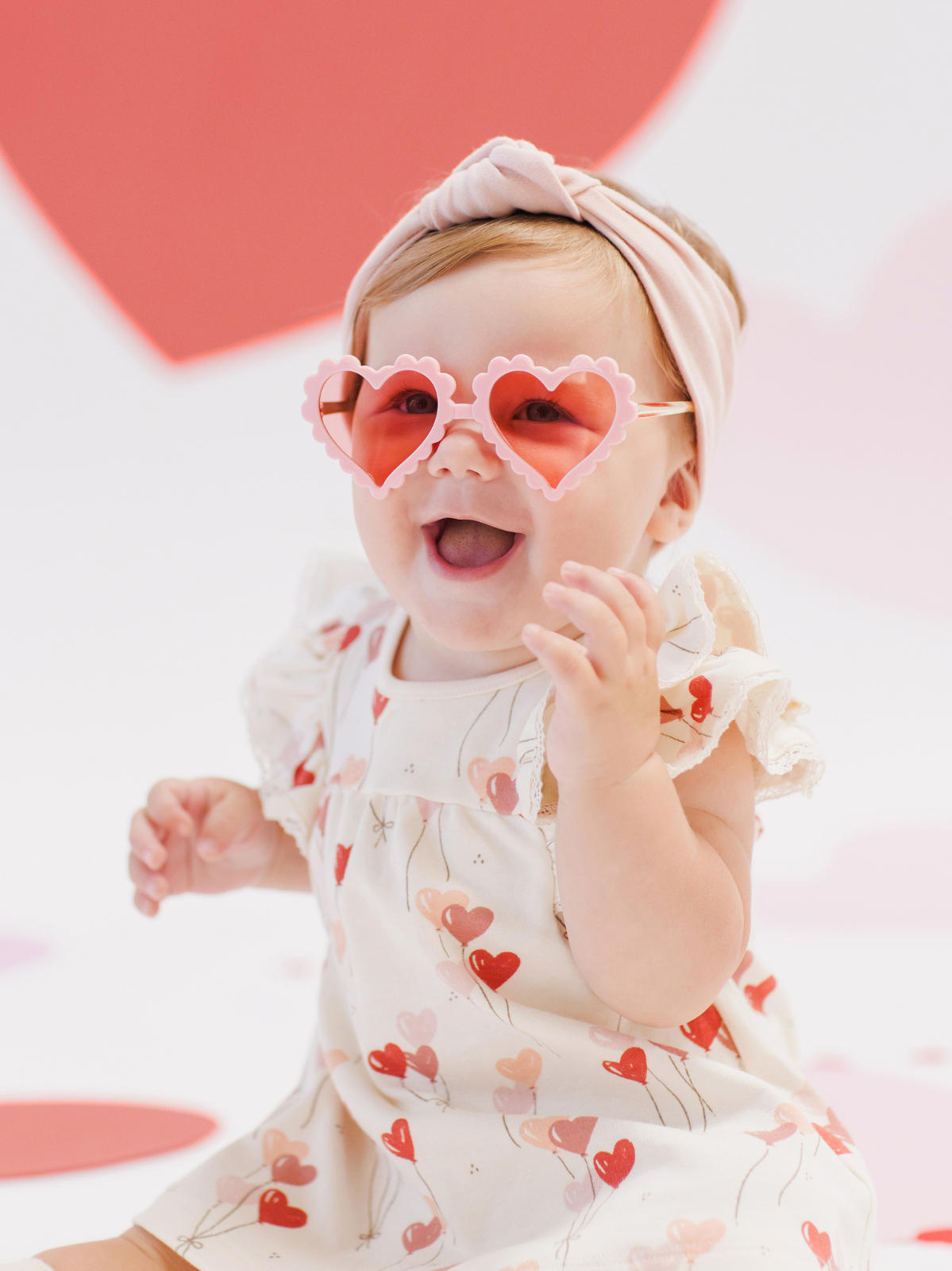 Smiling baby in heart-shaped sunglasses, wearing a floral dress, against a playful heart-themed background.