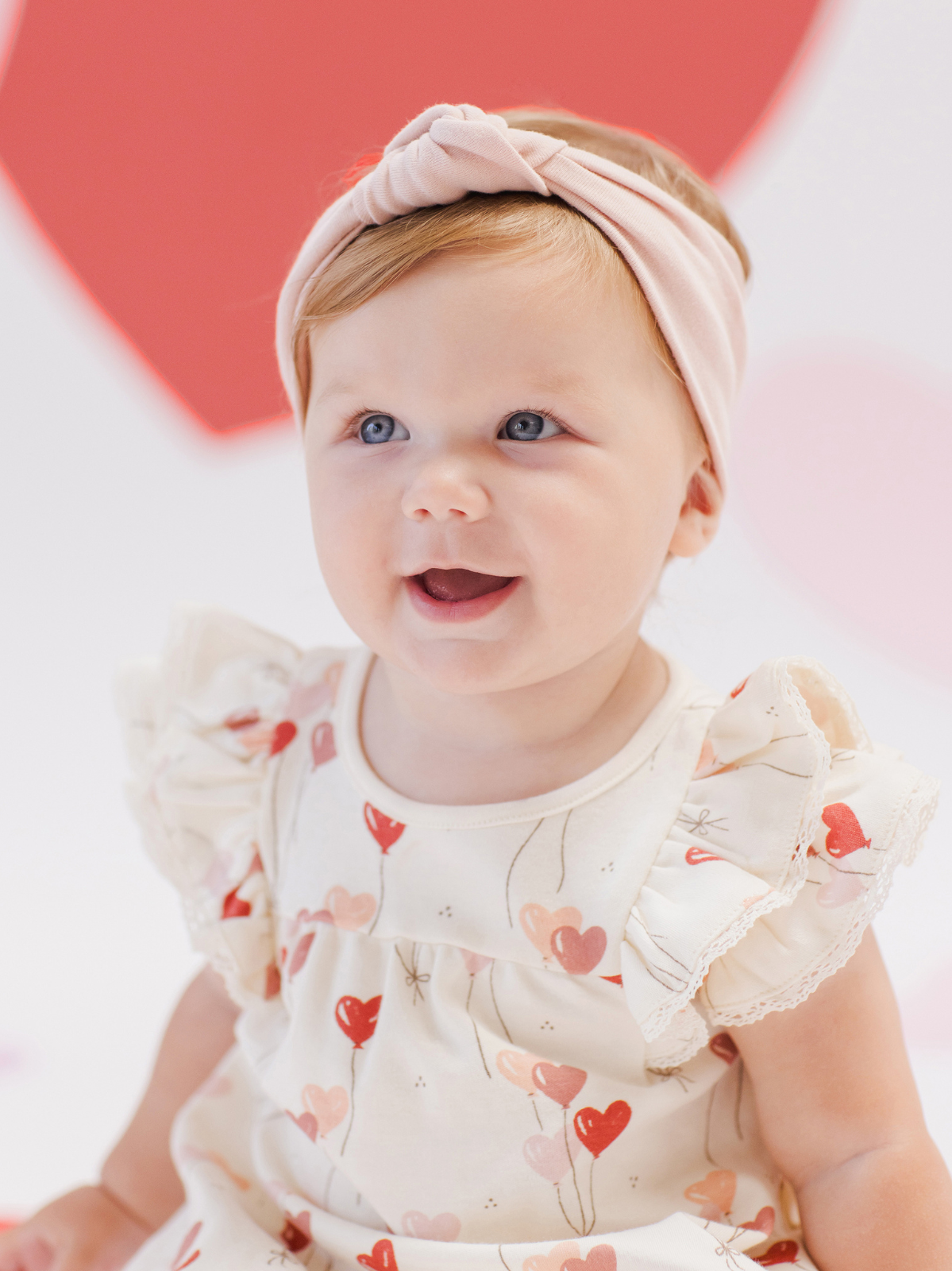 Smiling baby girl in a heart-patterned dress with a pink headband against a soft, colorful background.