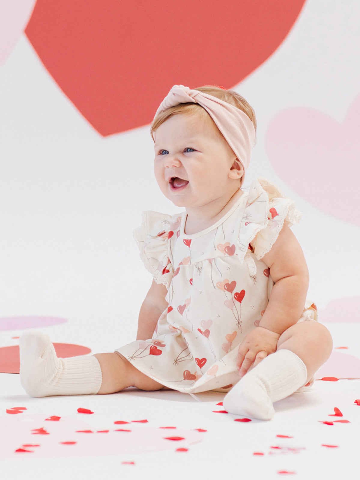 Baby girl in a heart-patterned dress sits on the floor surrounded by heart-shaped decorations, smiling joyfully.