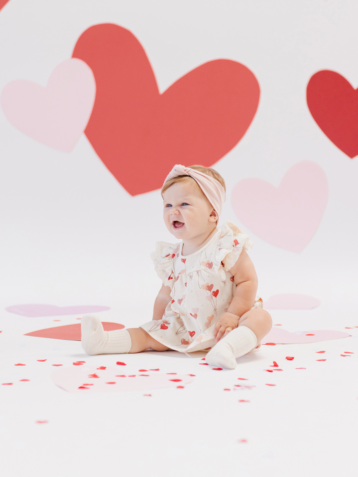 Baby sitting on a white background with heart decorations, wearing a heart-patterned dress and a pink headband.