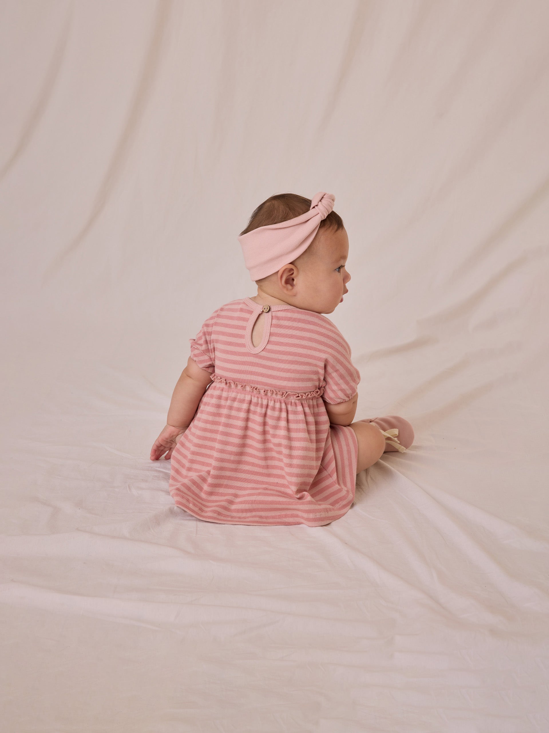 Baby in a pink striped dress and headband, sitting on a soft surface, looking away from the camera.