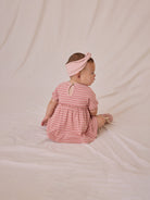 Baby in a pink striped dress and headband, sitting on a soft surface, looking away from the camera.
