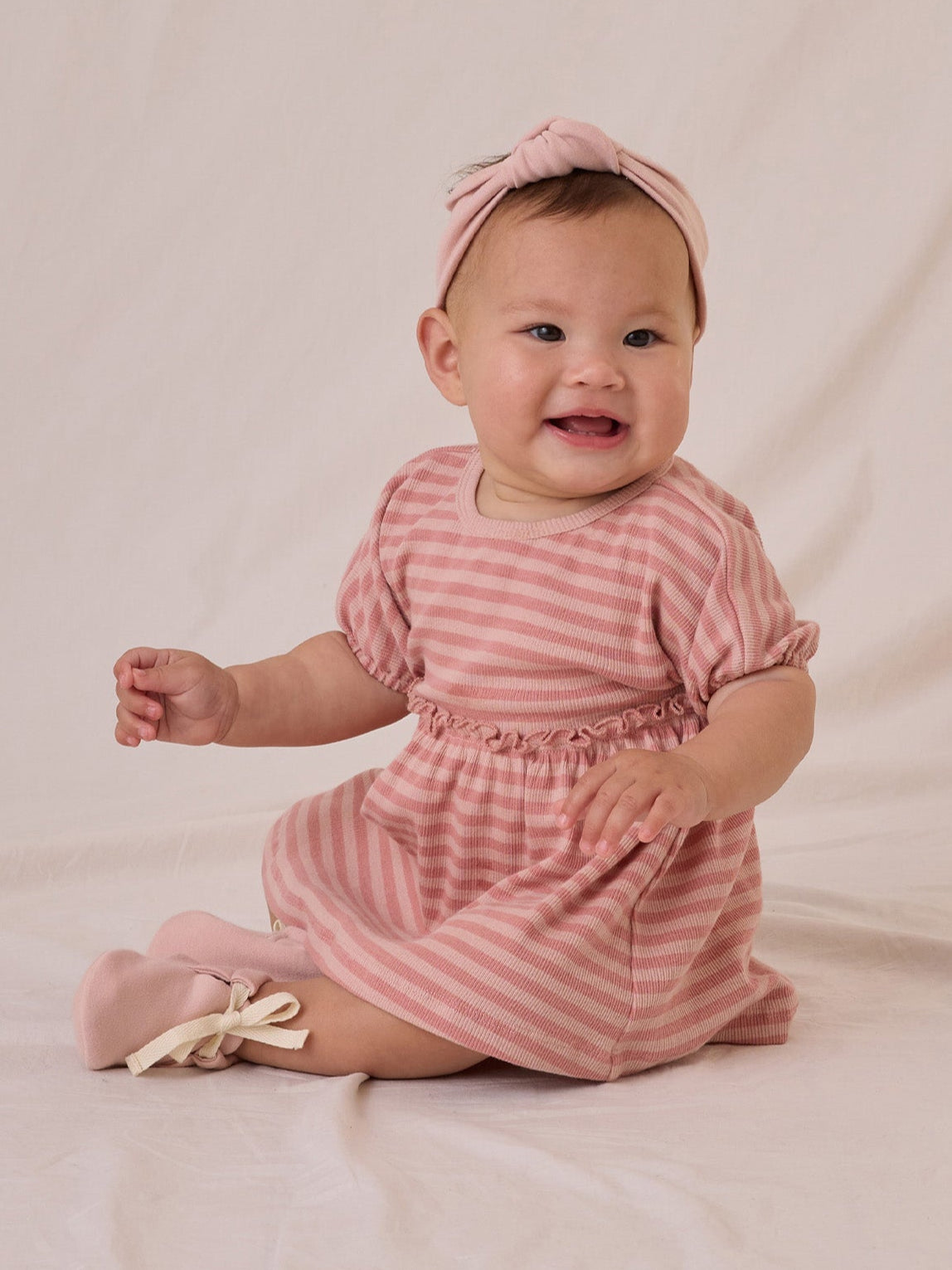Smiling baby in a pink striped dress and headband, sitting on a light background.