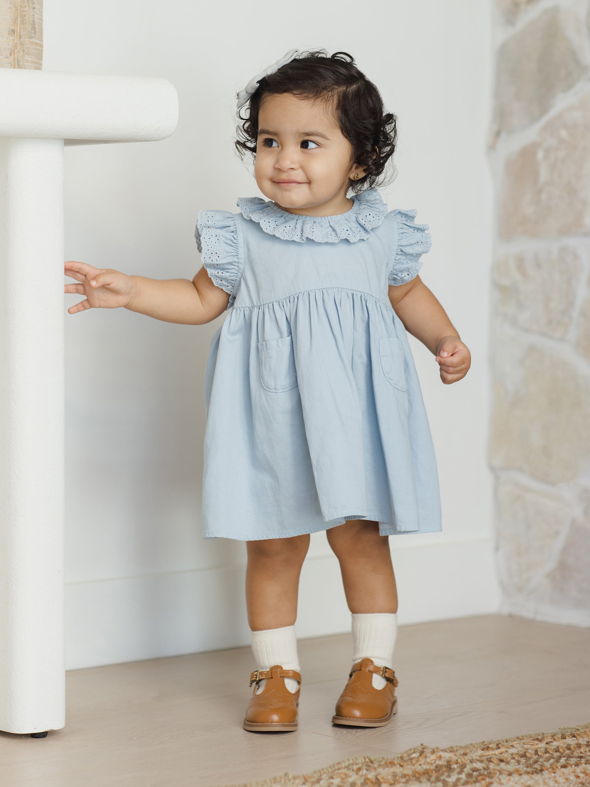 Smiling toddler in a light blue dress and brown shoes, posing by a wall with a stone accent.