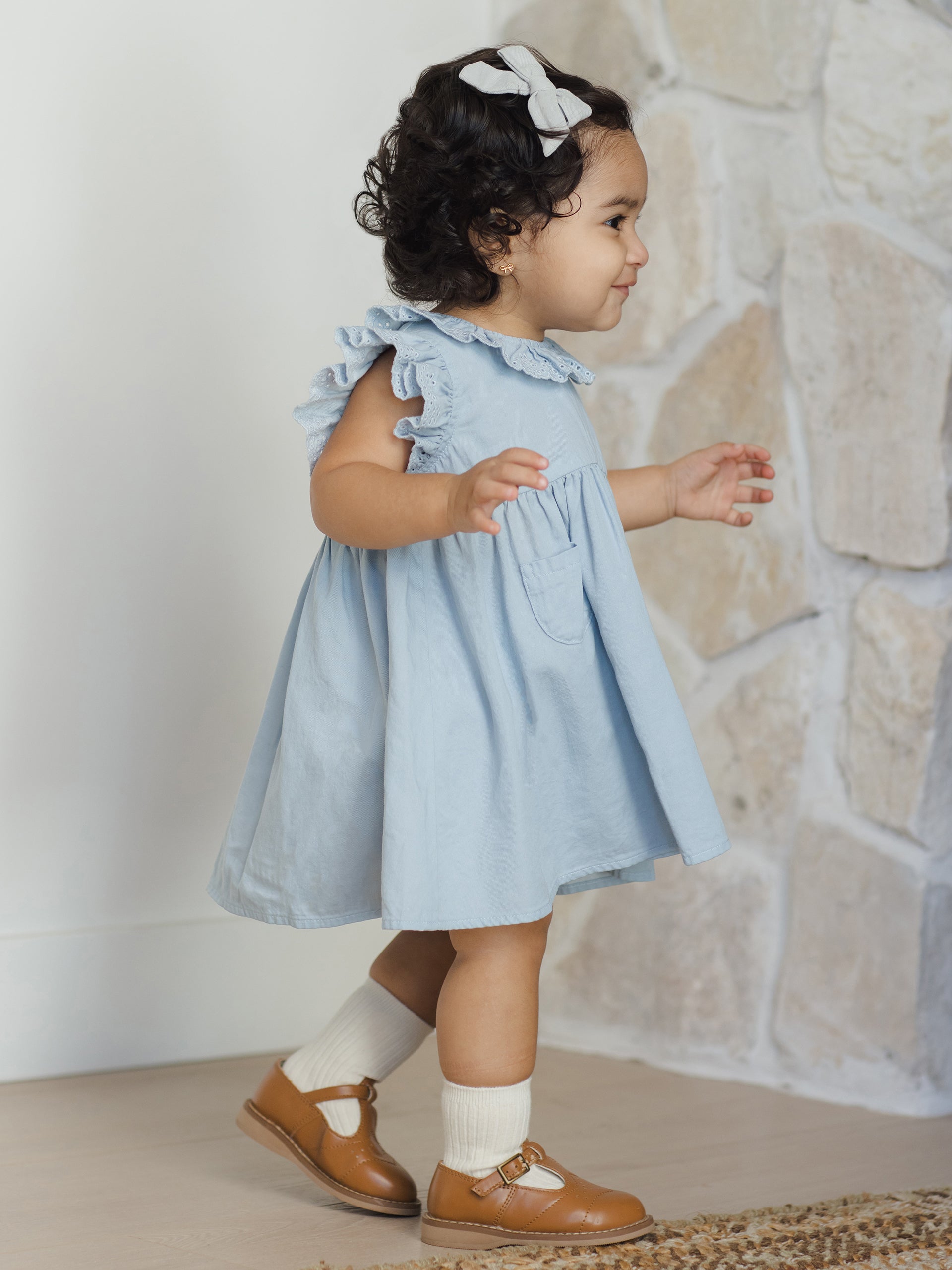 Toddler girl in a blue dress and brown shoes, smiling while walking near a stone wall.